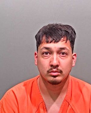 A man in an orange prison jumpsuit poses for a mugshot against a gray background.
