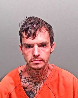 A man with tattoos on his neck and face is wearing an orange prison jumpsuit, looking directly at the camera.