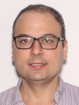 A man with glasses and short hair smiles at the camera, wearing a patterned shirt against a plain background.