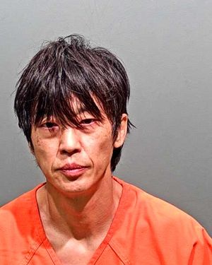 A man with messy hair is shown in a mugshot wearing an orange prison jumpsuit against a gray background.