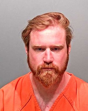 A man with a beard and disheveled hair is wearing an orange prison jumpsuit and gazing directly at the camera.