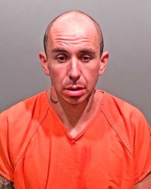 A man with a shaved head and tattoos, wearing an orange prison jumpsuit, appears to be looking downwards.