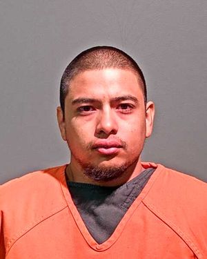A man with short hair and a beard is wearing an orange jail uniform and looking directly at the camera.