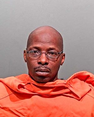 A man wearing an orange prison jumpsuit and glasses is photographed against a neutral background.