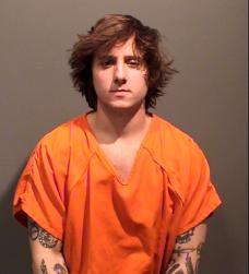 A person with tousled hair and tattoos is wearing an orange inmate jumpsuit and posing for a mugshot.