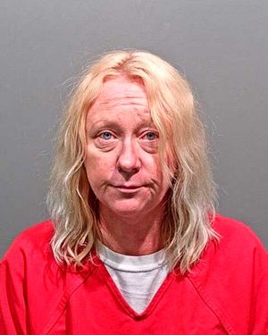 A person with long, light-colored hair is posing for a mugshot while wearing a red jail uniform.