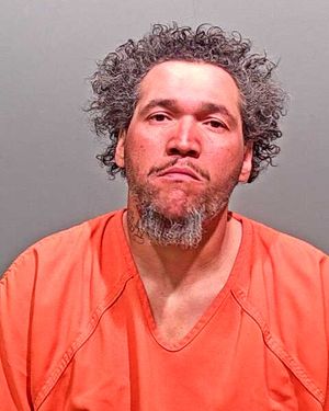 A man with curly hair and facial hair wearing an orange prison jumpsuit, looking directly at the camera.