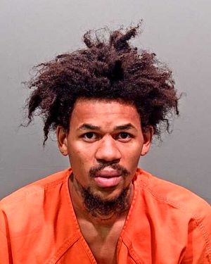 A man with unruly hair is shown in an orange prison jumpsuit against a gray backdrop.