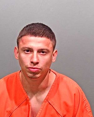 A person wearing an orange jail jumpsuit poses for a mugshot against a gray background.