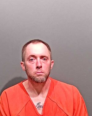 A man wearing an orange prison jumpsuit poses for a mugshot against a gray background.