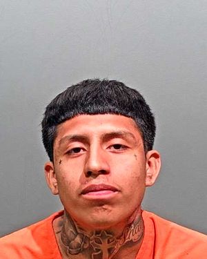 A young man with tattoos, wearing an orange shirt, is facing the camera against a plain gray background.
