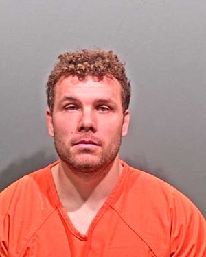 A man with curly hair is shown wearing an orange prison jumpsuit against a gray background.