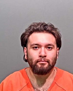 A man with a beard is shown wearing an orange prison jumpsuit against a gray background.