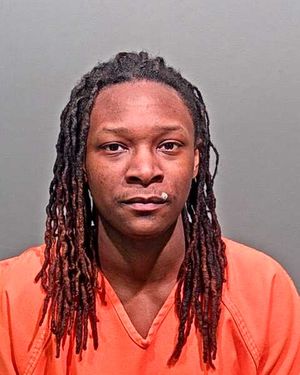 A person with long, braided hair is shown wearing an orange jumpsuit against a gray background.