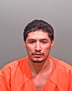 A man with short dark hair is shown in a mugshot wearing an orange prison jumpsuit against a gray background.