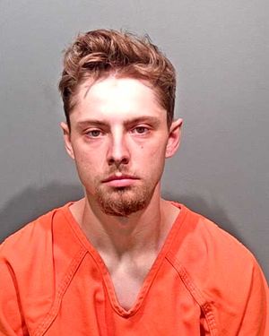 A male wearing an orange prison jumpsuit poses for a mugshot against a gray background.