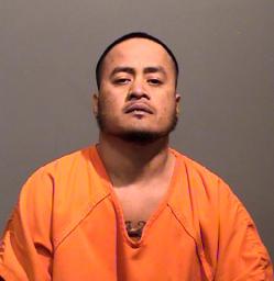 A man wearing an orange prison jumpsuit poses for a mugshot against a gray background.
