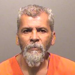 This image shows a man with a gray beard and hair, wearing an orange prison jumpsuit.