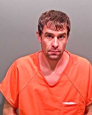 A man with short brown hair wearing an orange prison jumpsuit poses for a mugshot against a gray background.