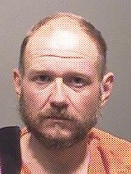 A man with a beard and shaved head is shown in a mugshot wearing an orange jumpsuit.