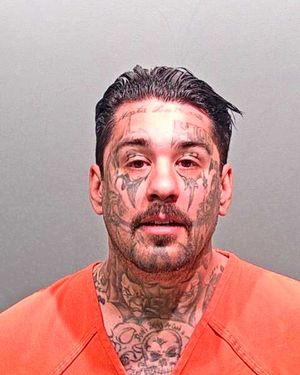 A man with extensive facial tattoos is wearing an orange jail jumpsuit and looking directly at the camera.