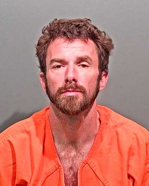 A man with a beard and tousled hair is shown wearing an orange prison jumpsuit and looking directly at the camera.