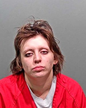 A woman with disheveled hair is shown in a police mugshot wearing a red jacket and appearing fatigued.