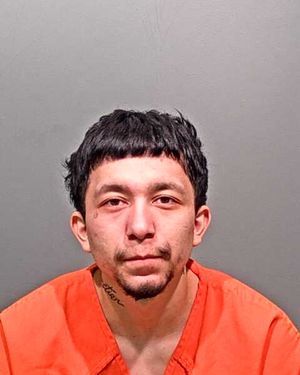 A young man with short black hair is wearing an orange prison jumpsuit and is posed for a mugshot against a gray backdrop.