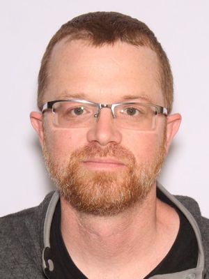 A man with glasses and a beard, wearing a gray jacket over a black shirt, poses for a portrait against a plain background.