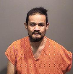 A man wearing an orange jumpsuit is seated against a plain background, looking directly at the camera.
