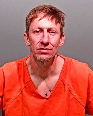 A man with short hair and a goatee is shown wearing an orange jumpsuit against a gray background.