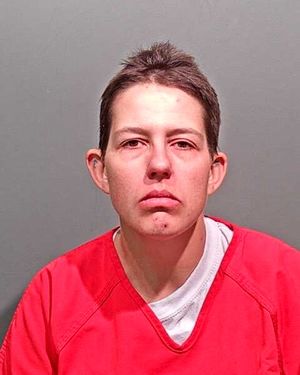 A person with short hair wearing a red prison jumpsuit looks directly at the camera against a plain gray background.
