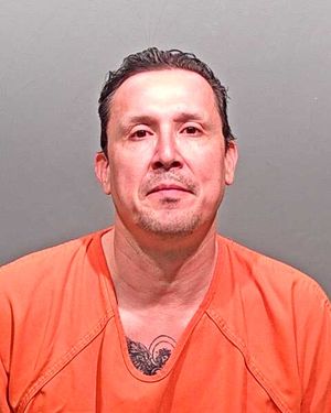 A man wearing an orange prison jumpsuit poses for a mugshot against a gray background.