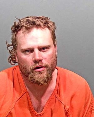 A man with a disheveled appearance and a beard is shown wearing an orange prison jumpsuit against a gray background.