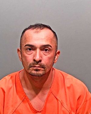 A man with a shaved head and a serious expression is wearing an orange prison uniform against a gray background.