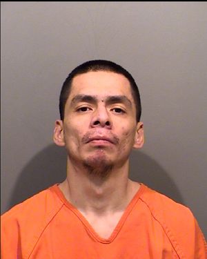 A man in an orange prison jumpsuit poses for a mugshot against a gray background.