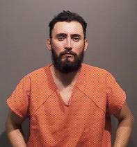 A man with a beard is wearing an orange prison jumpsuit and is posing for a mugshot against a plain background.