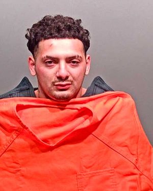 A young man with curly hair and facial marks is wearing an orange prison jumpsuit and looking directly at the camera.