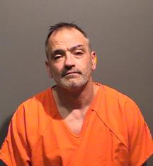 A man wearing an orange prison jumpsuit poses for a mugshot against a gray background.