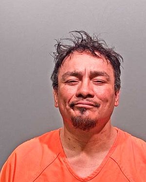 A close-up mugshot of a man with messy hair wearing an orange jumpsuit, smiling slightly.
