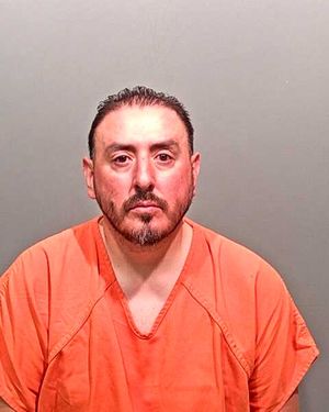 A man wearing an orange prison jumpsuit is posing for a mugshot against a gray background.