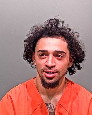 A man with curly hair and a beard is wearing an orange jail uniform and appears to be smiling in a mugshot.