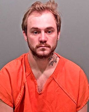 A man with a beard and tattoos is shown wearing an orange jumpsuit against a gray backdrop.