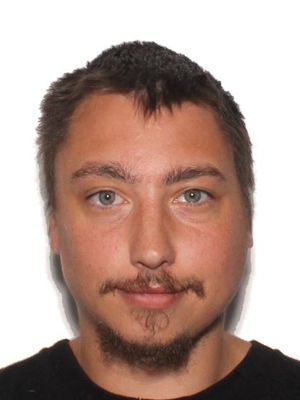 A close-up portrait of a young man with short hair, blue eyes, and facial hair, wearing a black shirt.