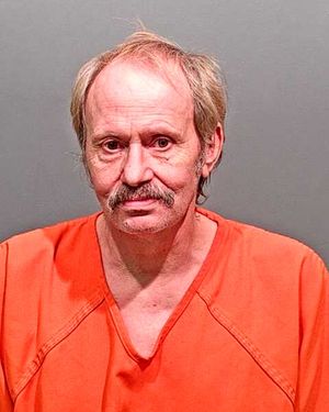 A man with a mustache is shown wearing an orange prison uniform against a gray background.