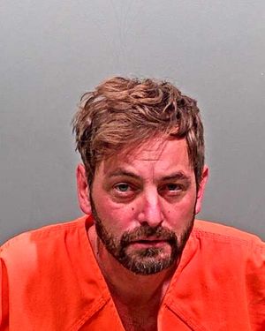 A man with a beard and disheveled hair wearing an orange jumpsuit is shown against a gray background.