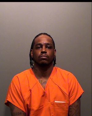 A man in an orange prison jumpsuit poses for a mugshot against a gray background.