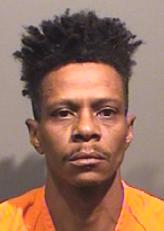 A man with short, curly hair is shown in a mugshot, wearing an orange prison jumpsuit.