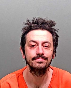 A man with messy hair and facial hair is wearing an orange jail jumpsuit and looking directly at the camera.