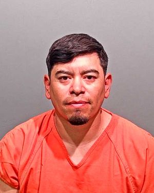 A man wearing an orange prison jumpsuit poses for a mugshot against a gray background.